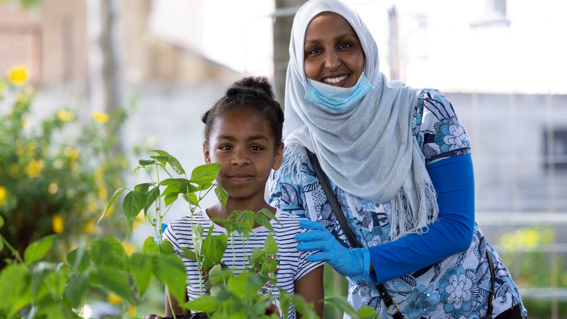 Woman wearing a headscarf with a child standing behind plants and smiling