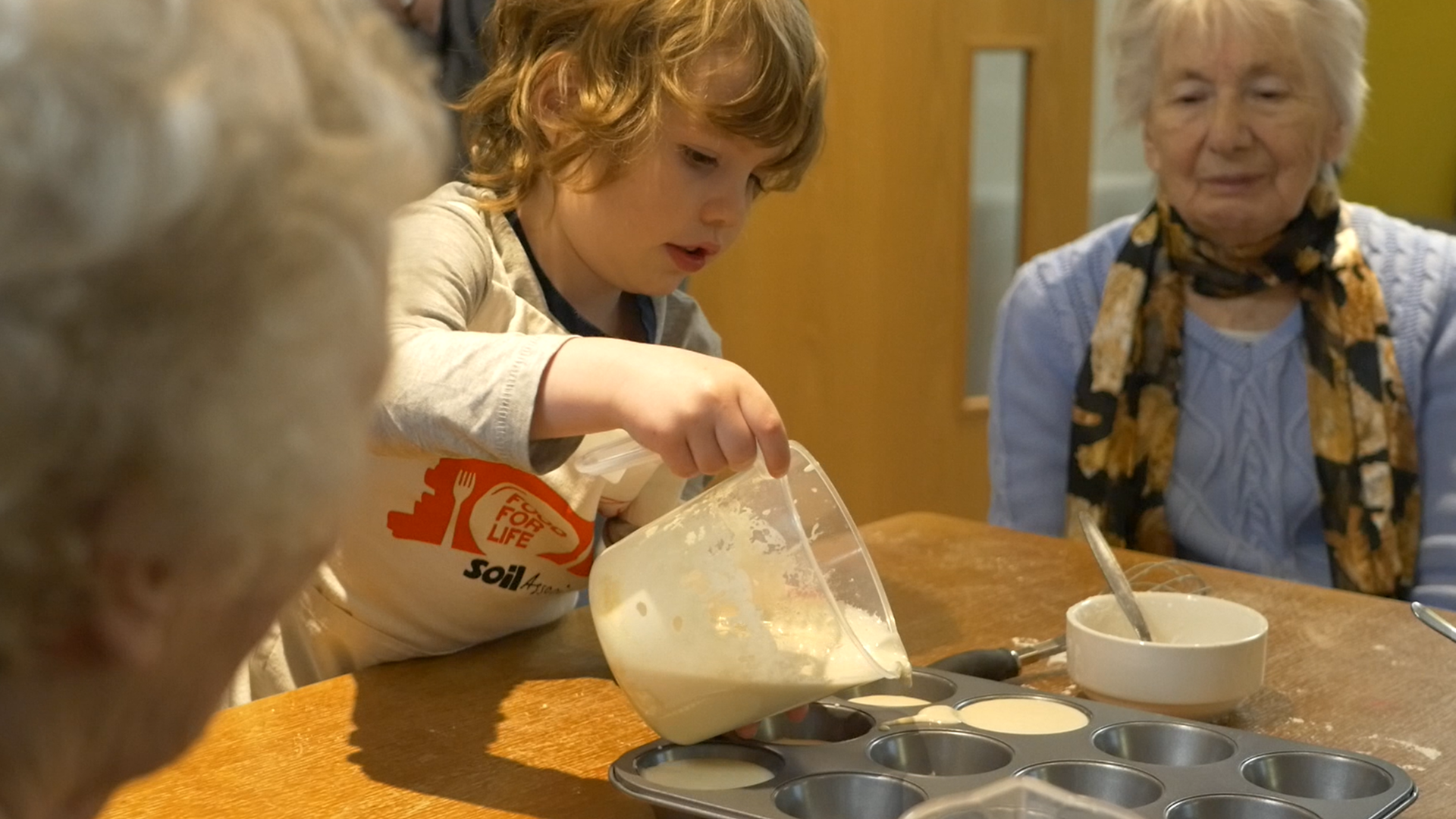 Older woman looks on as an early years child pours batter into a baking tray